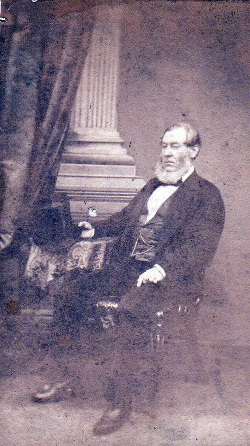 Faded carte de visite of a seated bearded man in a dark coat and waistcoat beside a column