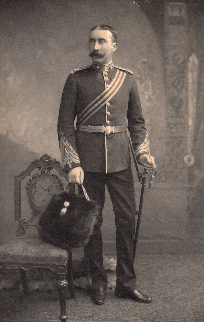 Studio photograph of a moustached officer in full dress uniform with sword, holding a fur cap