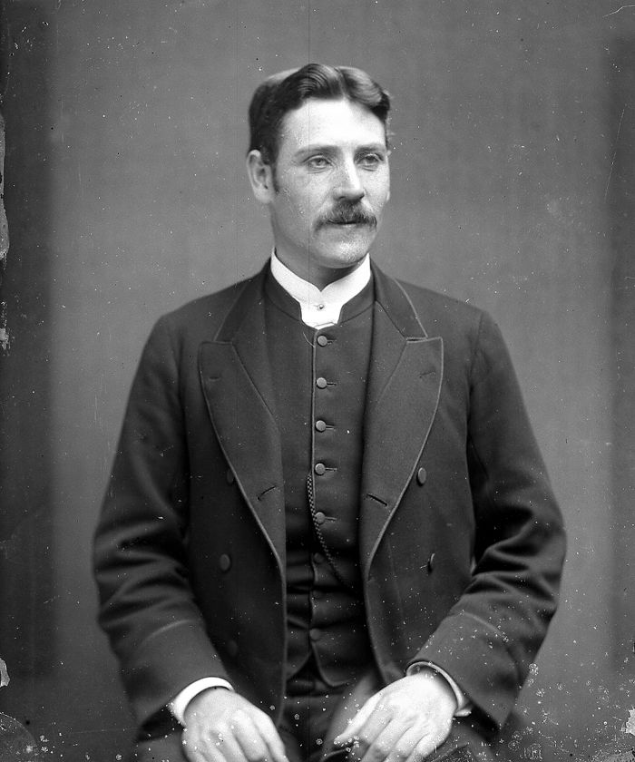Studio photograph of a young clergyman with a moustache, in a clerical collar and long double-breasted coat